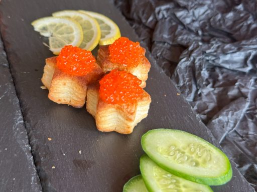 Lachs-Tatar mit Forellenroggen, Zitrone und Gurkenscheiben auf einem schwarzen Teller.