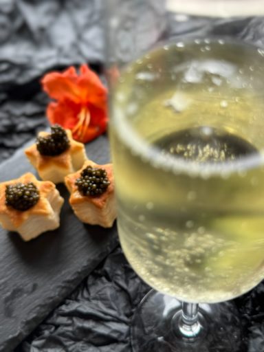 Glas mit sprudelndem Getränk und Häppchen mit Kaviar auf einem schwarzen Teller.